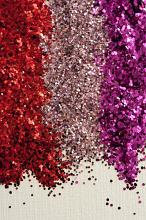 Colorful festive piles of red, pink and purple glitter for craft work arranged in three vertical lines over a textured white background with copy space