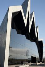 Modern exterior facade of the Glasgow Riverside Museum with its distinctive zigzag design above a large glass window reflecting a tall ship exhibited in the harbour