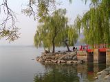 Tranquil Scenic View of Small Red Footbridge on Rocky Shore with Weeping Willow Trees by Lake on Calm Day with Water Reflection