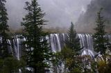 Misty waterfalls plunging over a mountain cliff in an ethereal landscape of steep forested slopes