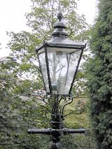 Old cast iron Victorian gas lamp with the traditional lantern style glass on a street with leafy green trees, close up view of the streetlight
