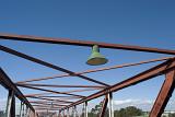 Overhead rusty girders of a bridge structure with an electric light with shade in an open framework against a blue sky