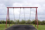 An empty swing set in a park with a view of the ocean on a cloudy day.