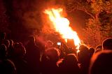 Crowd of people gathered around a bonfire with a large column of bright orange flames shooting into the air at night
