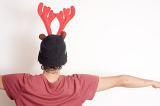 Young person wearing a Christmas hat with festive red reindeer antlers standing with back to the camera over a white wall and copy space