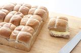 Scrumptious buttered hot cross bun on aa board with a knife and batch of fresh buns.