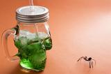 Creepy spider with a green Halloween drink in a glass skull shaped mason jar with lid and straw over an orange background with copy space