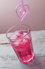 Close-up glass of red cocktail with ice cubes and curved heart-shaped pink straw, over old painted wooden surface