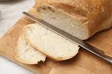 Slicing a loaf of fresh country bread off a round white cottage loaf with a bread knife on a wooden bread board