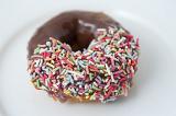 Chocolate glazed ring donut dipped in colorful candy sprinkles on one side on a white plate with focus to the sprinkles