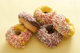 Pile of assorted fresh ring donuts or doughnuts glazed with chocolate, lemon or orange icing decorated with colorful sprinkles on a pale yellow background