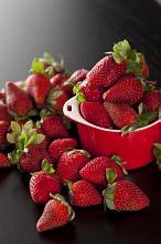 Juicy succulent ripe red strawberries with their green stems overflowing from a small red ramekin onto a dark background