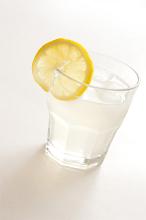Iced drink in a glass tumbler garnished with a slice of tangy lemon on white, clear liquid at a tilted angle