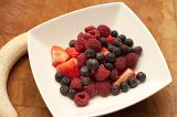 Bowl of fresh autumn fruit with assorted berries including strawberries, blueberries and raspberries with a peeled whole banana alongside the dish