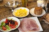 Cold buffet lunch laid out on a rustic table with pasta, sliced cured ham, cold pork pie and a mixed salad with olives