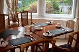 Dining table laid for breakfast with three place settings including coffee mugs beneath a window overlooking the garden