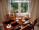 Stylish wooden dining table set for three people for breakfast with cutlery and crockery alongside a view window overlooking shrubs