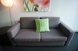 Upholstered grey settee with a single green cushion under a modern abstract picture illuminated by a standard lamp
