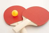 Two wooden red ping pong bats with a yellow ball lying on an off white surface viewed low angle