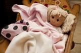 Blond toy doll wrapped in pink blankets lying in a wooden crib, close up view from above