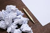 Difficult work and writers block concept with a bunch of crumpled papers and a clean sheet with metal pen on the surface of wooden table, viewed in close-up