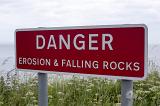 Erosion and falling rocks danger sign with white letters on red well visible background, installed outdoors near the sea