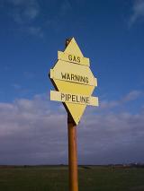 Gas pipeline warning sign in countryside with black letters on yellow background on the pole in the field. Viewed from low angle against cloudy sky