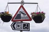 Quayside warning sign on a metal pole with hanging baskets of summer flowers and WC sign for handicapped people