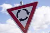 Warning sign for an approaching traffic roundabout in a red triangle against a cloudy blue sky