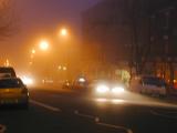 Cars driving at night in urban fog with headlamps and street lights illuminated against decreased visibility and dangerous driving conditions