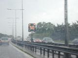 An illuminated 50 speed sign on a motorway in the center behind a safety barrier on a rainy day with traffic