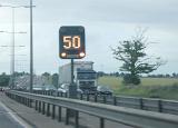Illuminated digital motorway traffic speed sign in the centre of a multi lane highway with trucks and cars on a rainy day