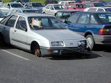 Old silver sedan car parked in a parking lot with a missing front tyre with a notice taped to its windscreen as the axle rests on the tarmac