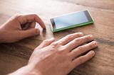 Close up of a mans hands as he taps on a wooden table anxiously while waiting for a call on a smart phone.