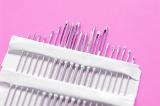 Close up on set of various sized sewing needle points in paper package organized by height and thickness over pink background