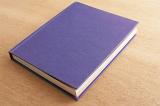 Closed hardcover notebook with a purple cover lying diagonally on a wooden desk in a close up view with copy space