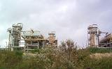 Infrastructure of an industrial plant or factory with external pipework viewed over bushes against a cloudy sky
