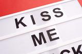 Close-up cropped image of white lightbox with black changeable sign Kiss Me, on red background. Love message concept