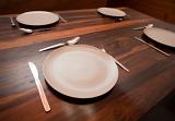 Empty white ceramic dinner plates and cutlery laid out on a bare wooden dining table