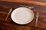 Place setting on bare wooden table with a plain white ceramic plate and cutlery, high angle close up view