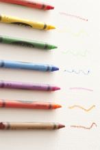 Row of colored wax crayons in the colors of the spectrum or rainbow with hand drawn squiggles showing the colors on white, overhead view
