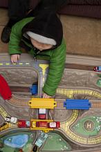 Top down view of young child in green winter coat and black hood playing with vehicles on large toy train and road set