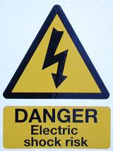 Large triangular yellow electrical warning sign with text below - Danger, Electric Shock Risk against a hazy blue sky