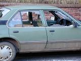 Old green car with smashed passenger windows form vandalism or an accident parked in front of a brick wall with shards of glass on the ground